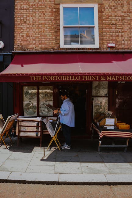 A person standing outside the Portobello Print & Map Shop under a red awning titled 'The Portobello Print & Map Shop' on a brick building facade. The individual is dressed in a light blue shirt, dark trousers, and a face mask, and is holding a clipboard or menu. Several wooden-framed tables with chairs are positioned on the sidewalk in front of the shop, some with paper or print materials on them. Next to the person, there are cardboard boxes and plastic containers stacked, indicating activities related to packing or receiving deliveries. The image is taken during daylight with clear weather, and a large window and a smaller window are visible above the shop, reflecting the sky and nearby structures. The scene suggests a typical street-level storefront, possibly part of a home relocation or moving process involving packing or material handling by North Kensington Removals.