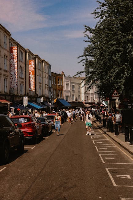 A busy street scene on Portobello Road in Ladbroke Grove showing pedestrians walking along the pavement and crossing the road, with parked cars lining both sides of the street. On the left side, a row of shops with colorful banners hangs above storefronts, and several people are seen standing and browsing. On the right side, black metal bollards separate the sidewalk from the parking area, where vehicles are parked closely together. In the foreground, a few cardboard boxes and plastic-wrapped furniture pieces are visible on the pavement near the entrance of a property, indicating ongoing home relocation or furniture transport activities typical of professional removals services. The sky is partly cloudy, and a large leafy tree extends over part of the scene, contributing to the urban street environment. The presence of moving-related objects and the context of loading or unloading suggests a residential move managed by North Kensington Removals during a busy day of packing and house removals.